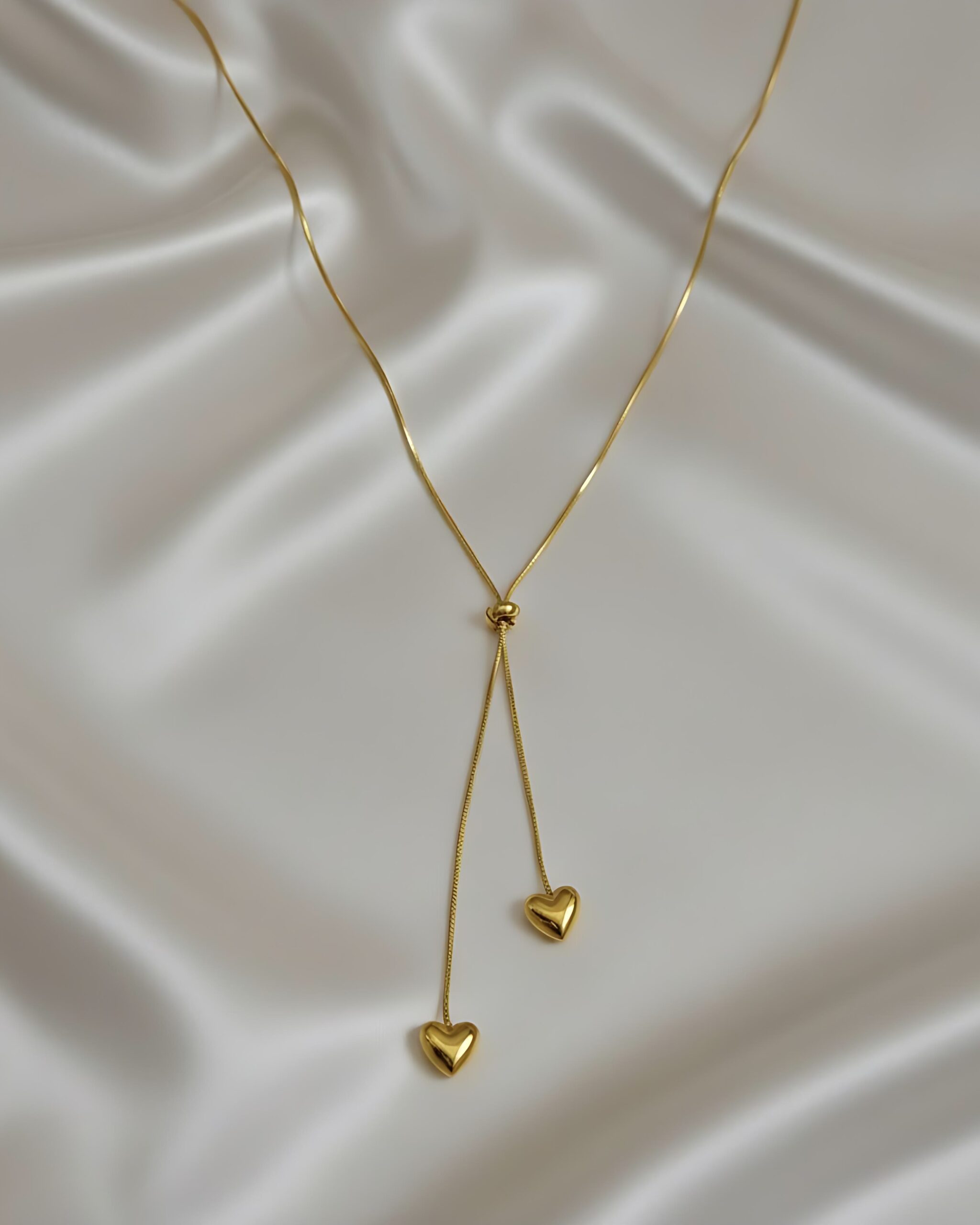 Sleek Gold Sleek Chain Drop Necklace with Puffy Heart Charms A gold lariat-style Y-necklace with a fixed drop design featuring a sleek snake chain and two dangling puffy heart charms at the ends, resting on a white silk background.