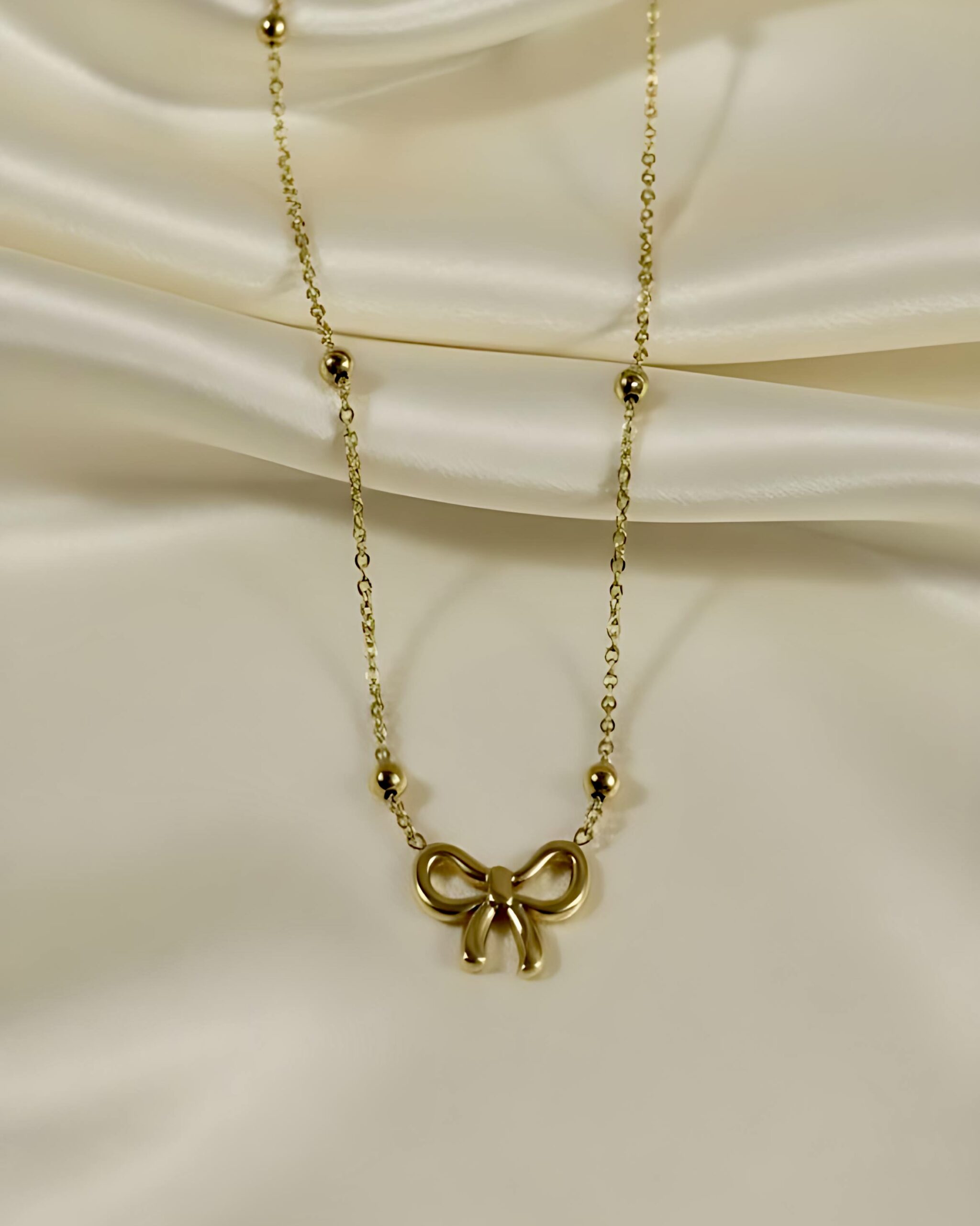 Gold Bow Necklace with Beaded Chain - Feminine Charm Jewellery A gold necklace featuring a dainty, polished ribbon bow charm pendant on a fine beaded or satellite chain, resting on a white silk background.