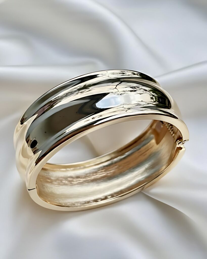 A wide, high-polished gold hinged bangle bracelet featuring a smooth, contoured or wave-like design, resting on a white silk background.