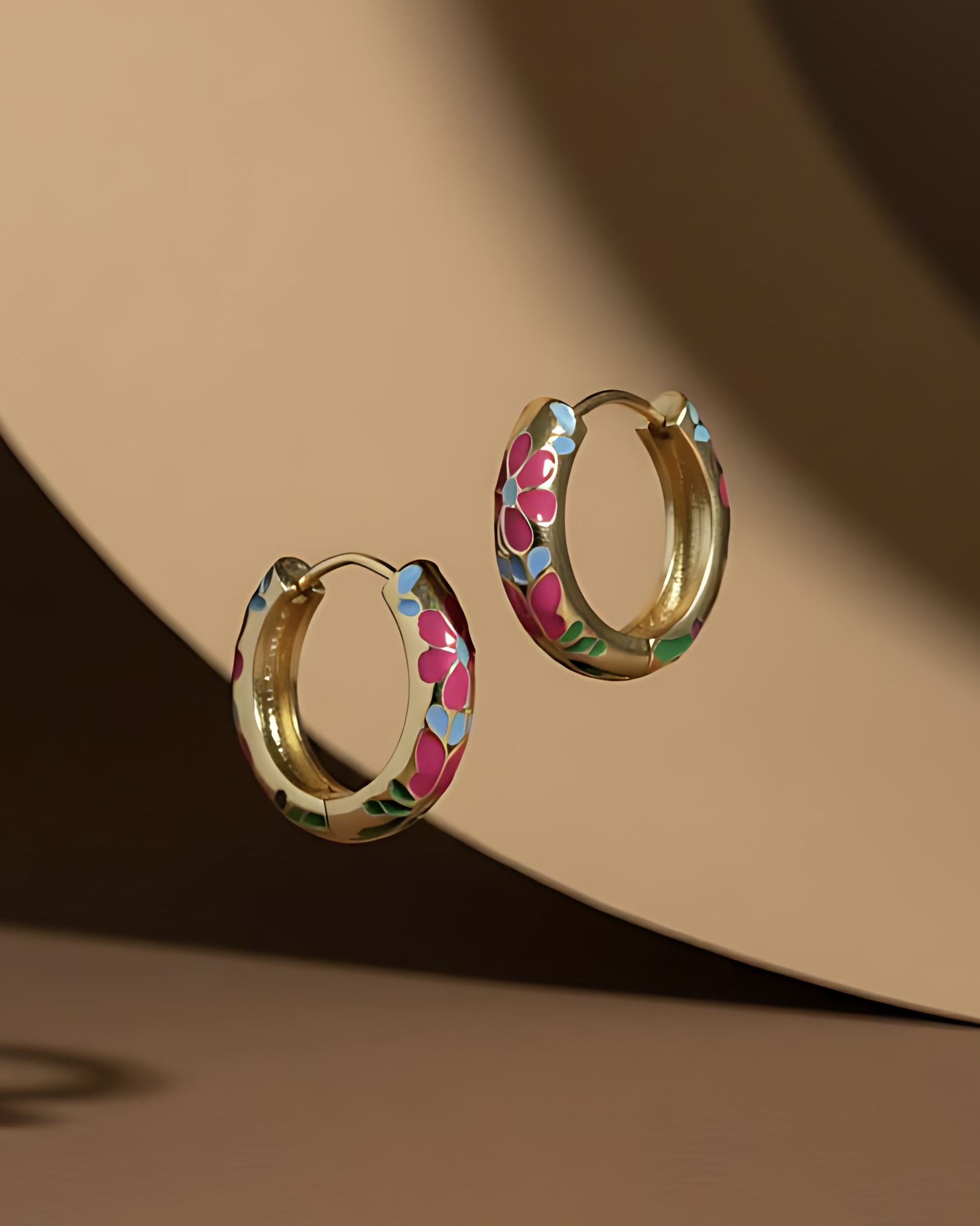 Hand-Painted Enamel Flower Hoop Earrings A pair of gold huggie hoop earrings featuring a continuous, hand-painted floral pattern in vibrant pink, blue, and green enamel, set against a brown background.