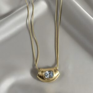 A gold necklace featuring a wide, chunky, retro-style pendant set with a central square white gemstone, suspended from a double snake chain on white silk.