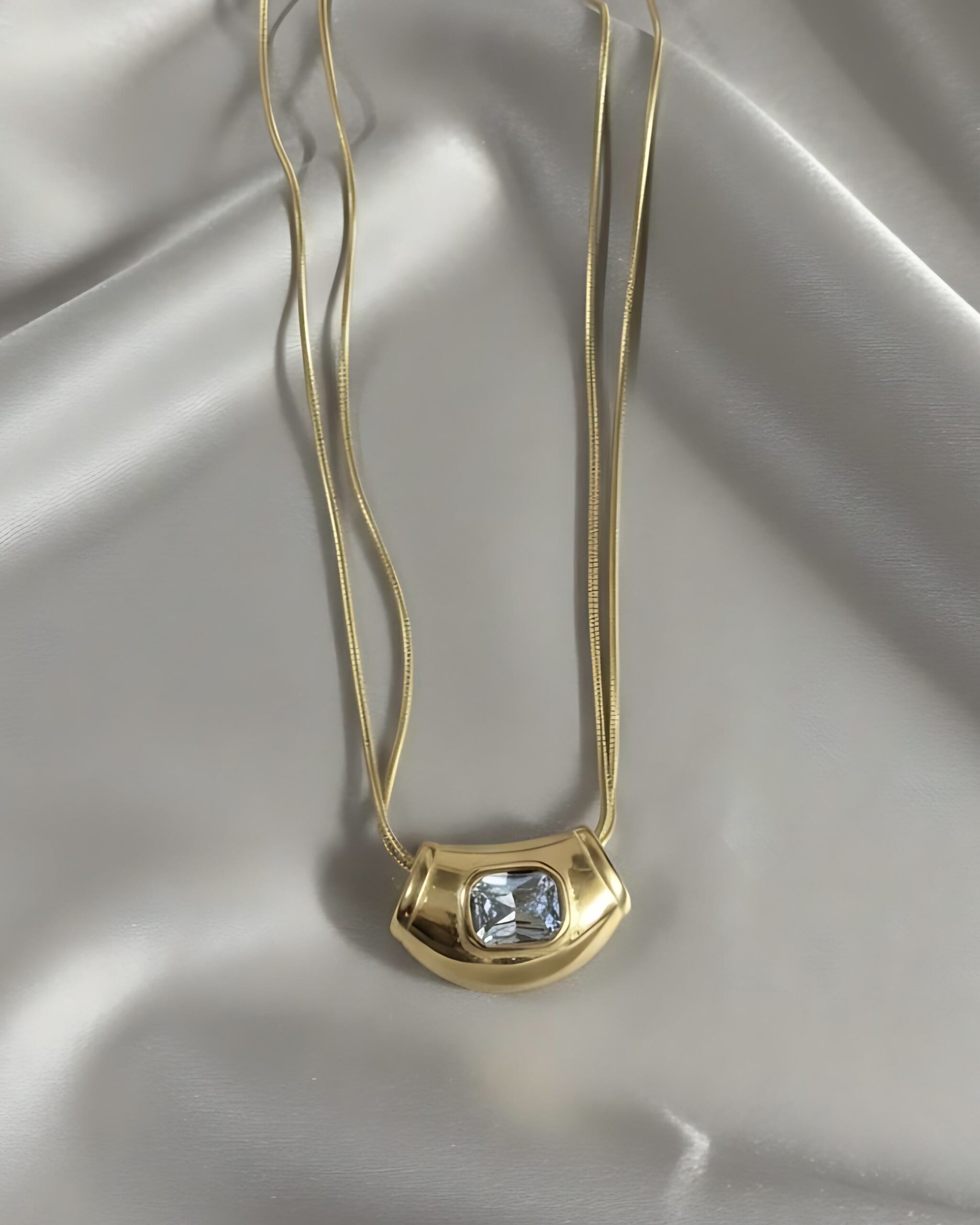Sleek Snake Chain Necklace with Vintage-Inspired Statement Jewellery A gold necklace featuring a wide, chunky, retro-style pendant set with a central square white gemstone, suspended from a double snake chain on white silk.