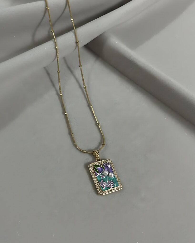 A gold satellite chain necklace with a small, rectangular pendant featuring a hand-painted floral enamel scene and a crystal border, resting on a gray surface.