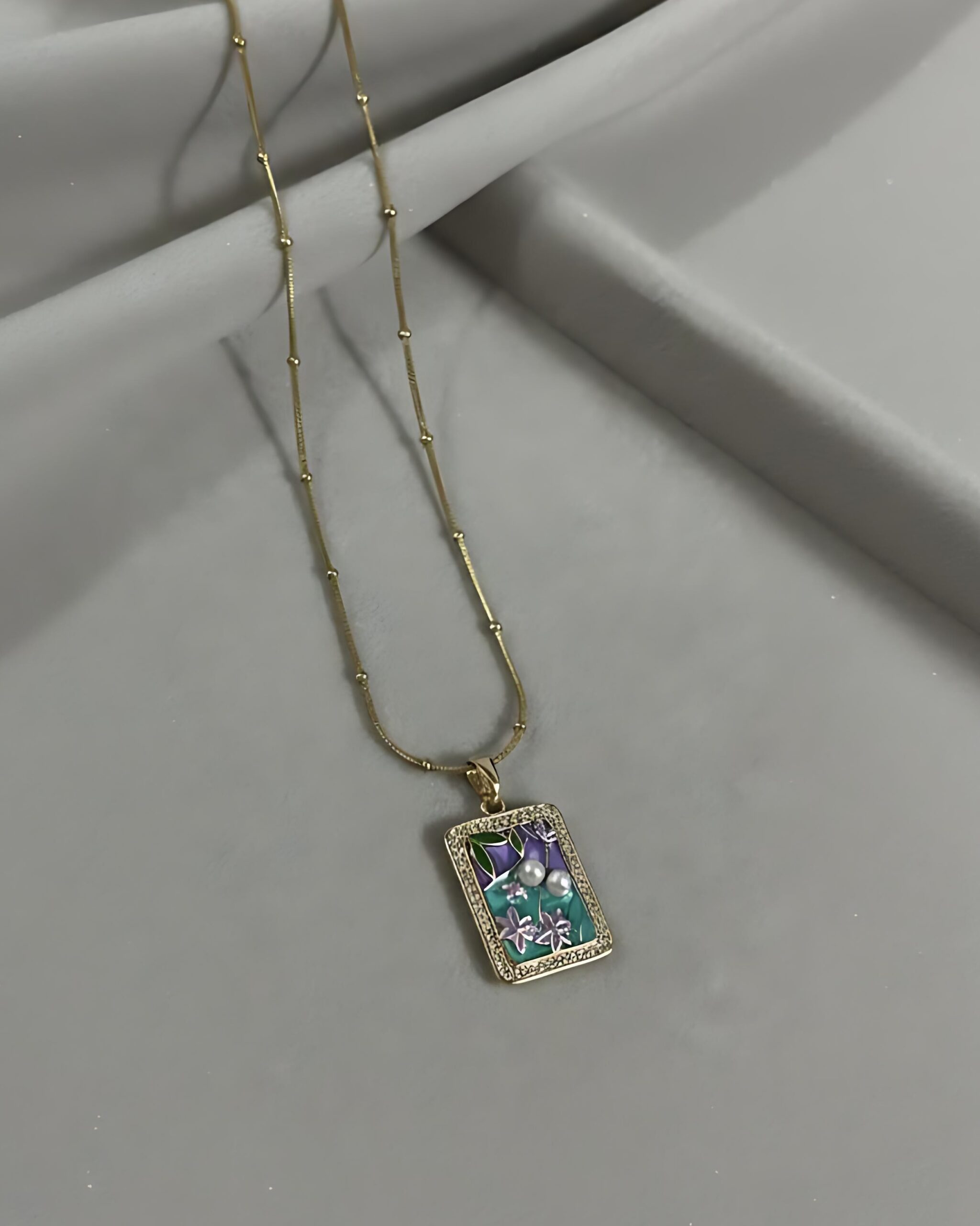 Gold Rectangle Pendant Necklace with Enamel Floral Art - Dainty Statement Jewellery A gold satellite chain necklace with a small, rectangular pendant featuring a hand-painted floral enamel scene and a crystal border, resting on a gray surface.