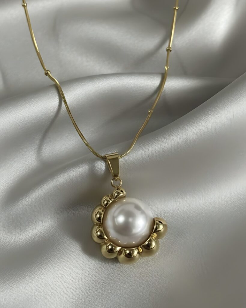 A gold necklace featuring a large, white pearl pendant framed by a chunky gold beaded sunburst design, suspended from a gold snake chain on white silk.
