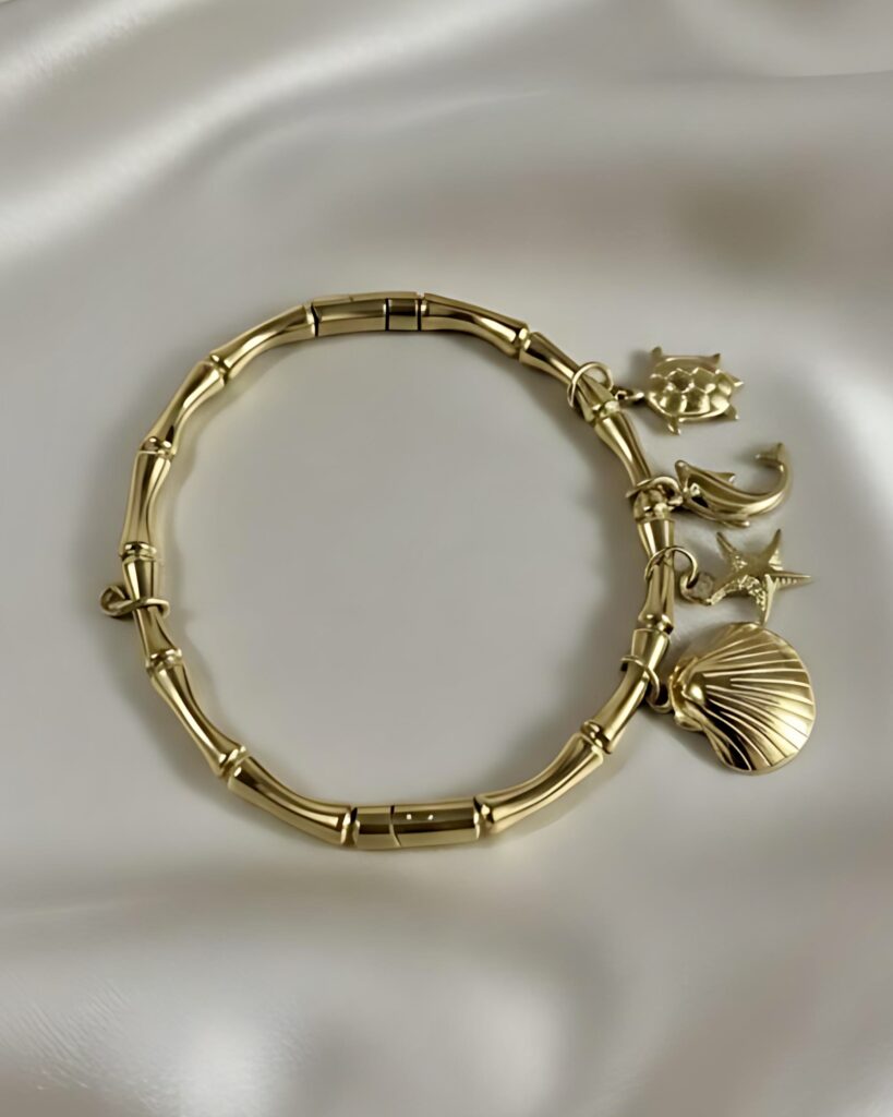 A gold bracelet with a thick, textured bamboo link band, adorned with four dangling ocean charms: a seashell, starfish, dolphin, and crab, resting on white silk.