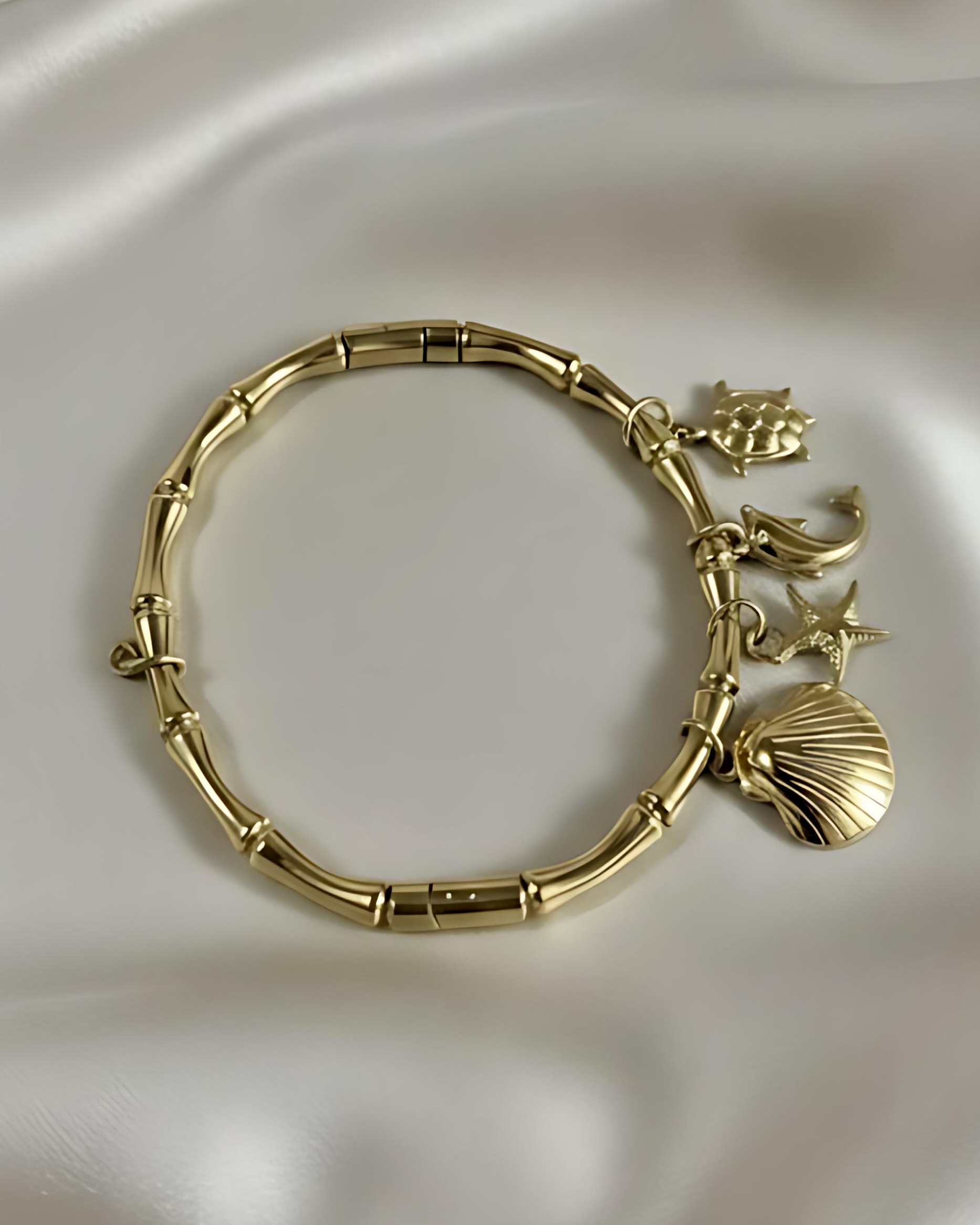 Gold Bamboo Link Charm Bracelet with Ocean Charms A gold bracelet with a thick, textured bamboo link band, adorned with four dangling ocean charms: a seashell, starfish, dolphin, and crab, resting on white silk.
