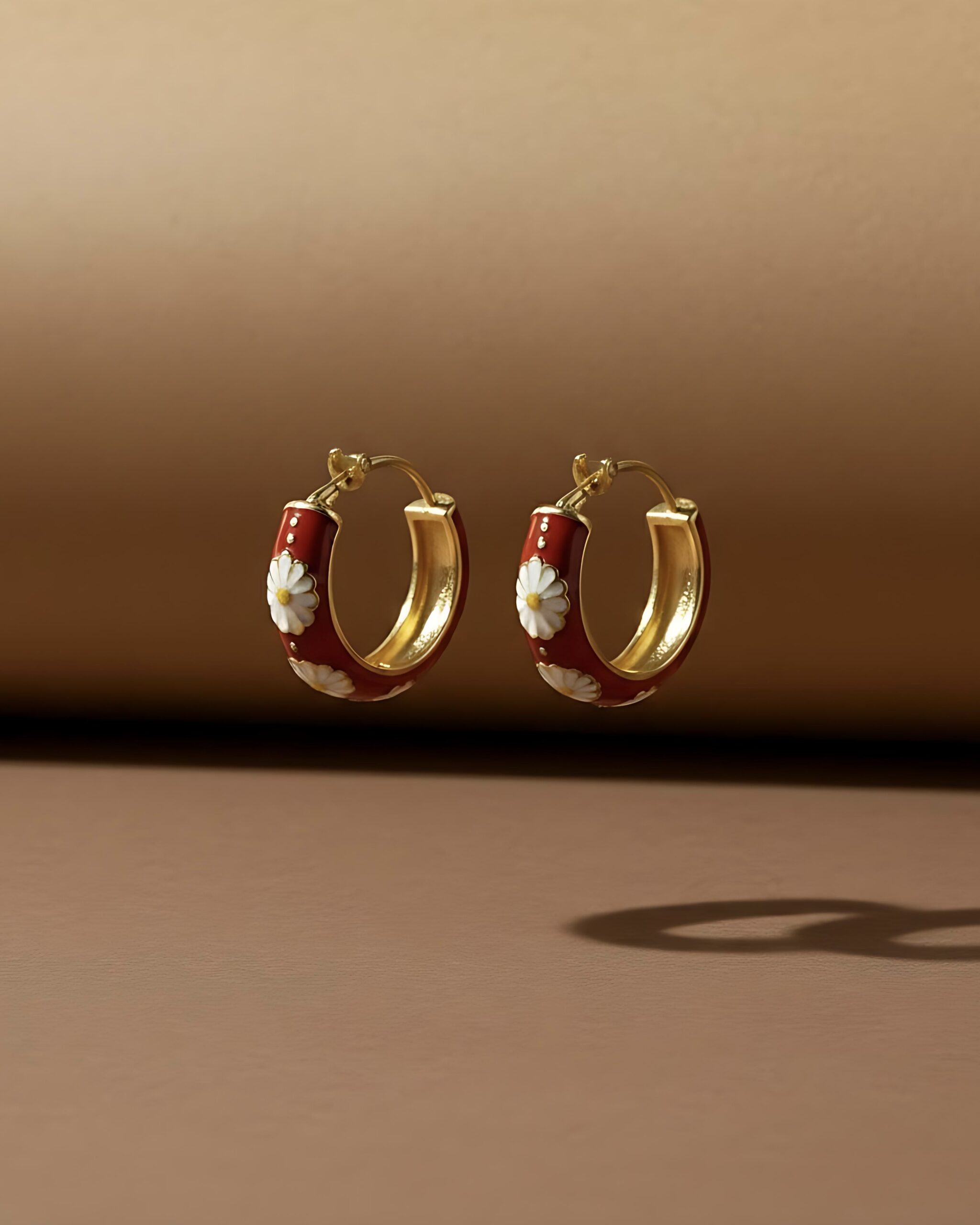 Gold Red Enamel Daisy Hoops- Bold Floral Statement Jewellery A pair of gold huggie hoop earrings featuring a continuous pattern of white daisy flowers over a bold red enamel background, set against a brown background.