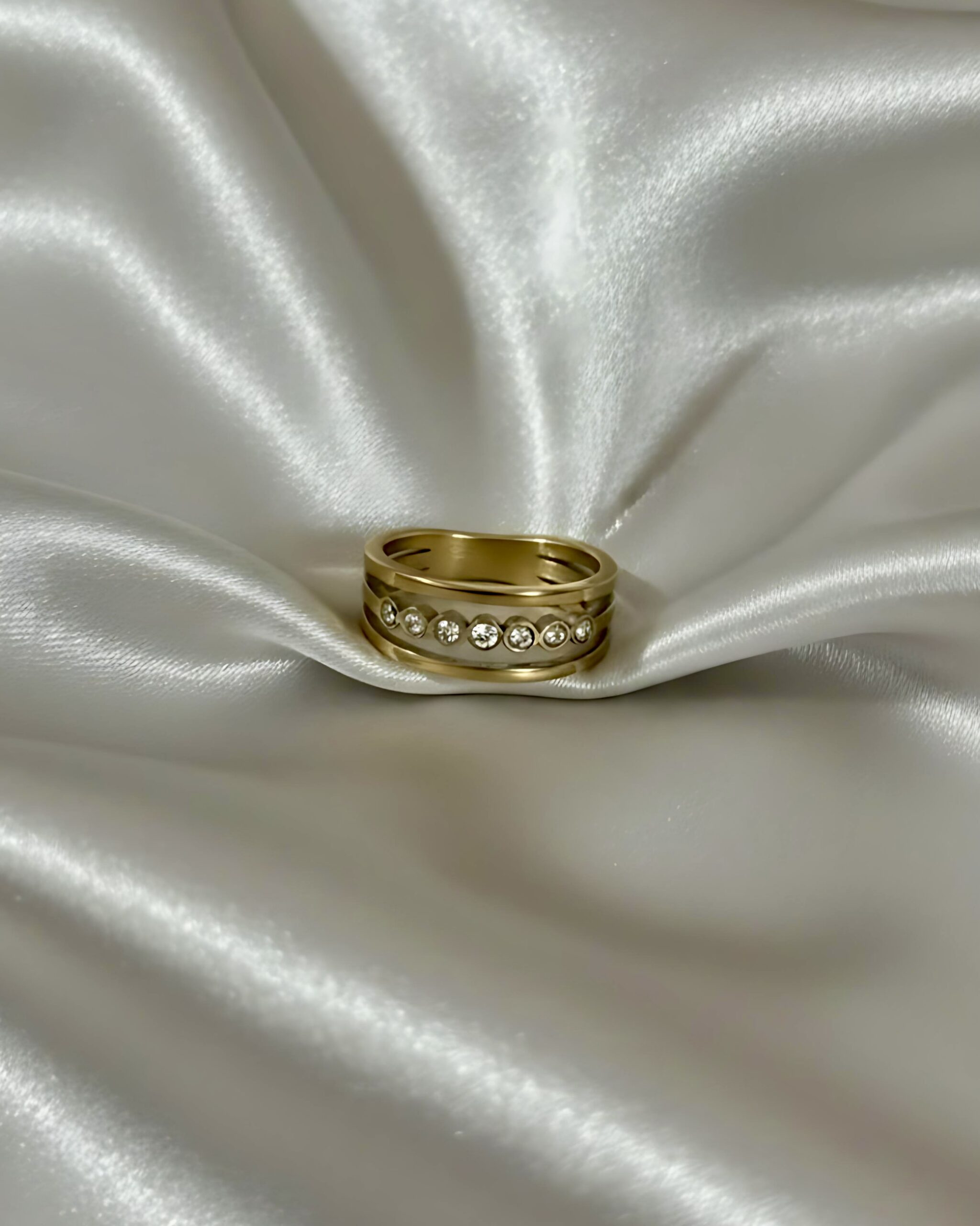 Five-Stone Crystal Band Ring in High-Polish Gold A wide gold band ring featuring a multi-layer design with a central channel of five round cubic zirconia crystals, resting on white silk.