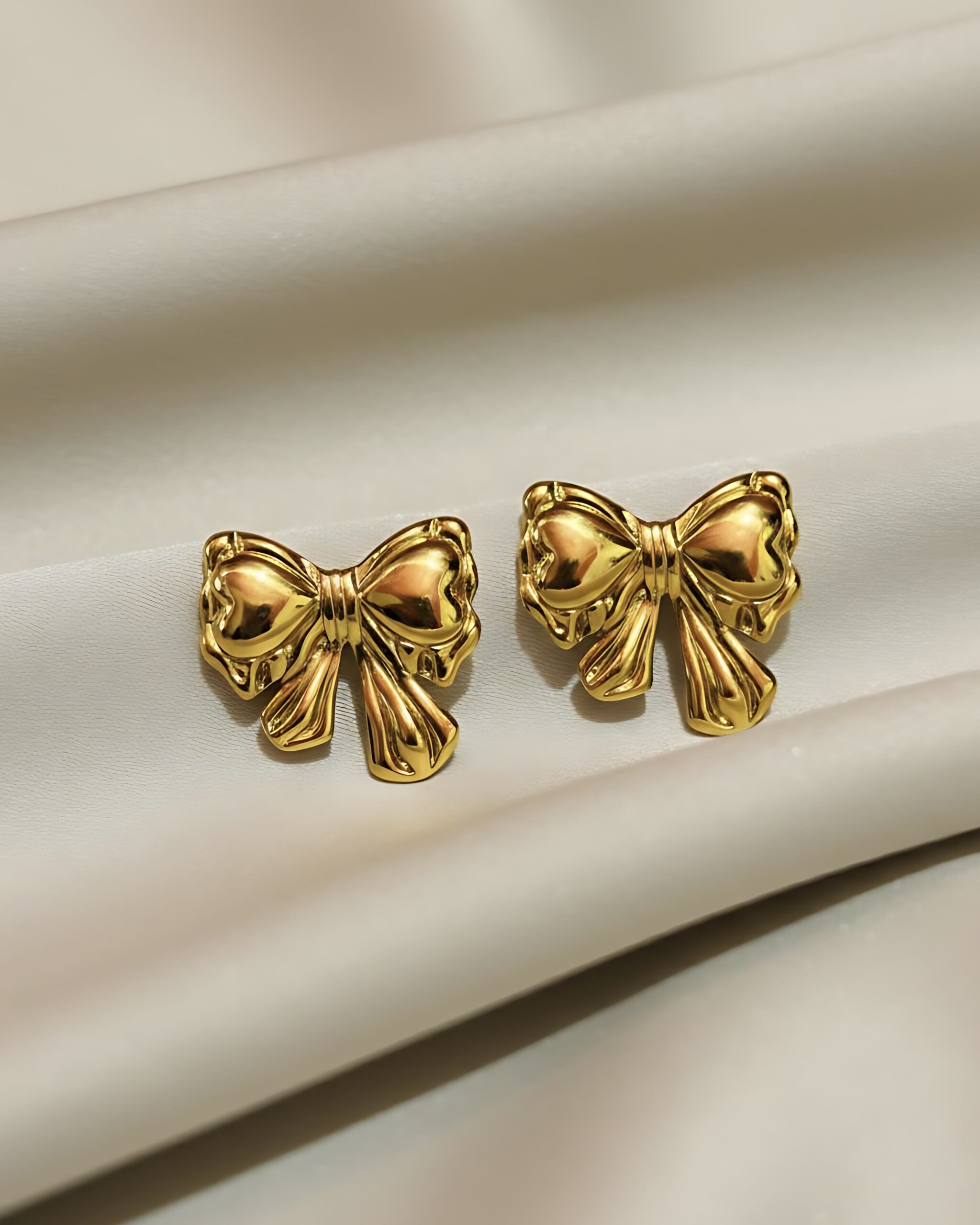 Minimalist Gold Bow Earrings-Everyday Feminine Jewellery A pair of gold stud earrings featuring a three-dimensional, puffy ribbon bowknot design with a high-polish finish, resting on white silk.