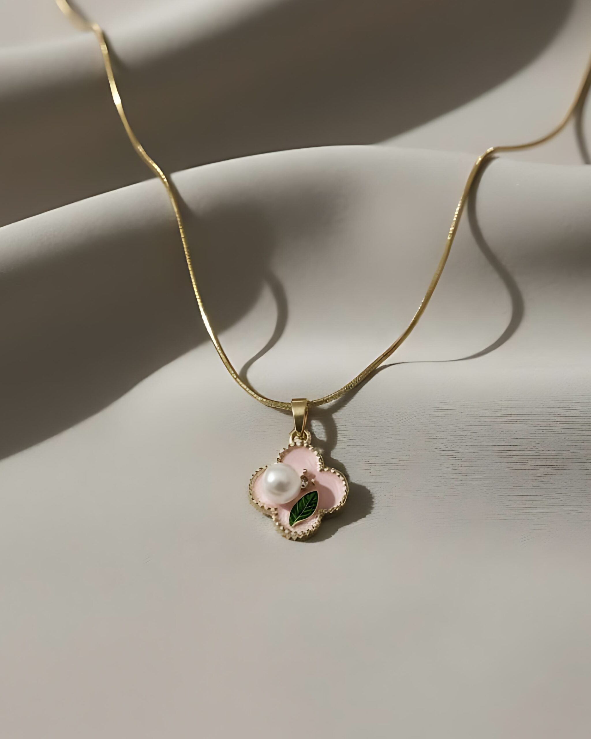 Gold Pink Enamel Clover Pendant Necklace With Pearl Accent A gold snake chain necklace with a small four-leaf clover pendant featuring pastel pink and green enamel with a small pearl center, resting on a gray and white background.