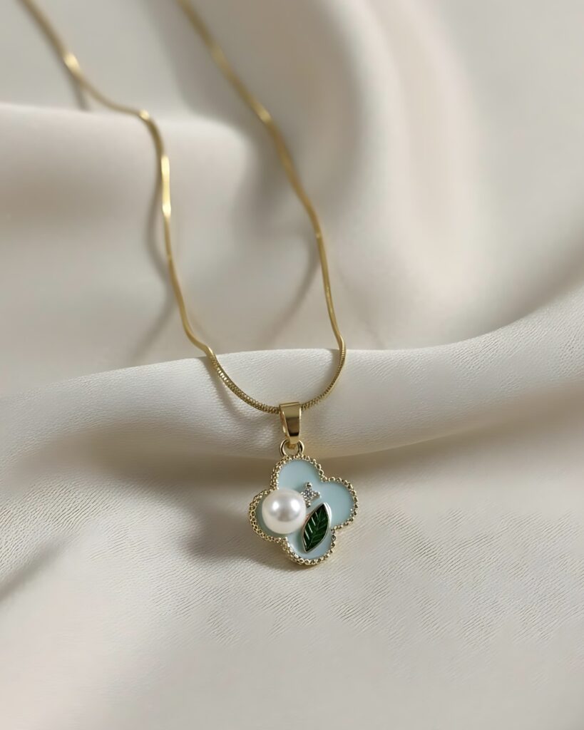 A gold snake chain necklace with a small four-leaf clover pendant featuring pastel blue and green enamel, a pearl accent, and a crystal pave border, resting on white silk.