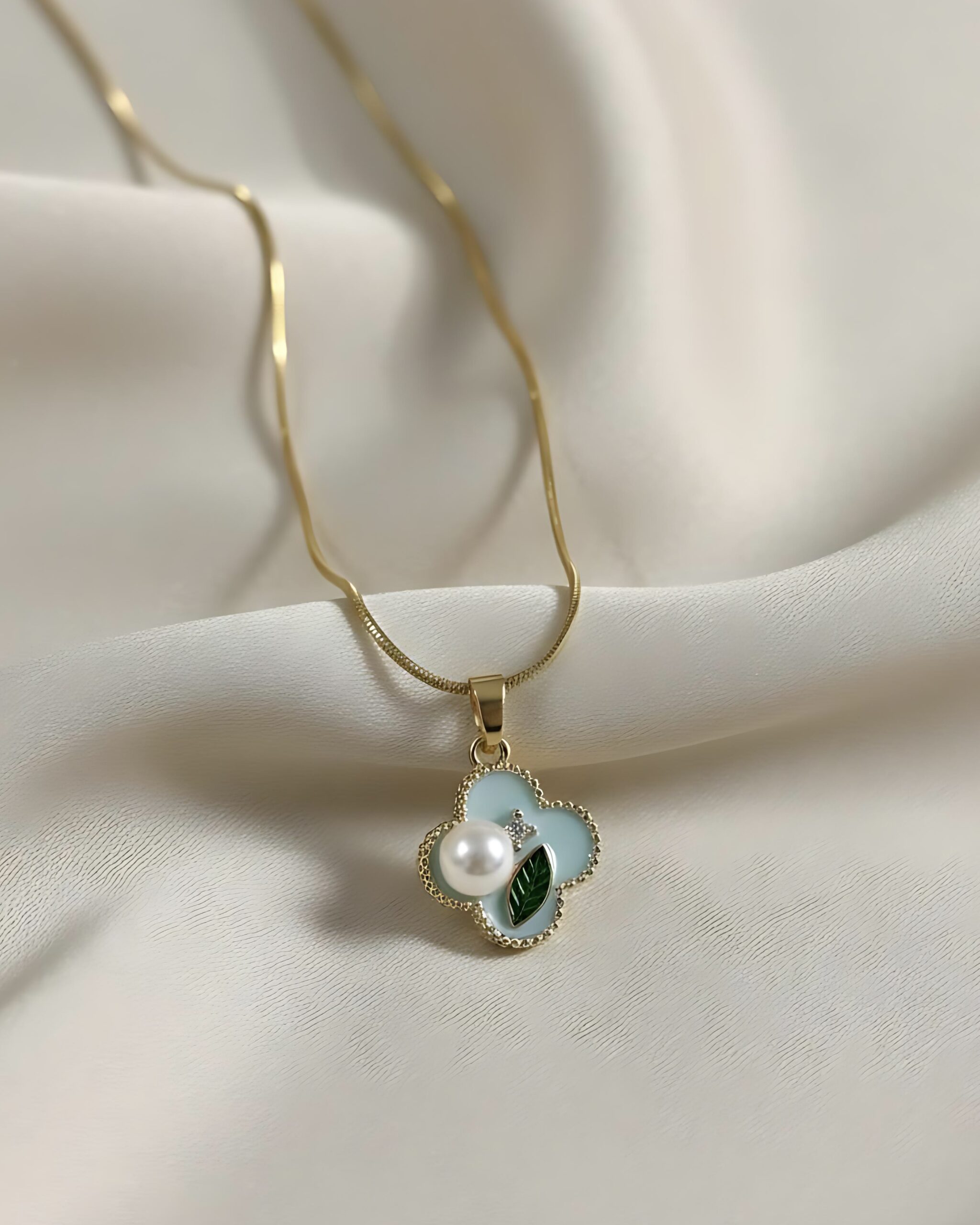 Gold Pastel Enamel Four - Leaf Clover Necklace with Pearl Accent A gold snake chain necklace with a small four-leaf clover pendant featuring pastel blue and green enamel, a pearl accent, and a crystal pave border, resting on white silk.