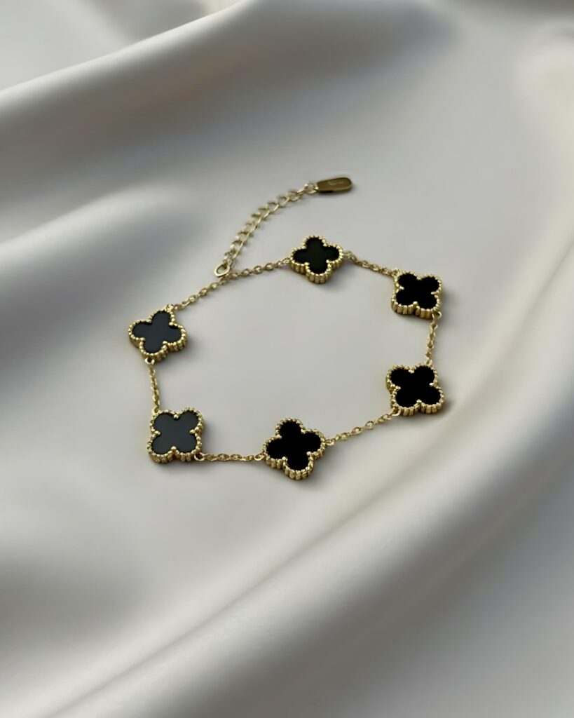 A gold link bracelet featuring five four-leaf clover charms. The charm visible is black enamel with a beaded gold border. This design is reversible to white mother-of-pearl.