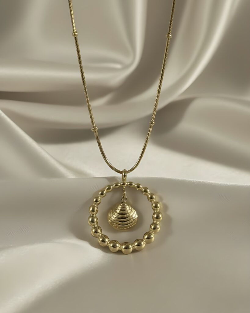 A gold necklace featuring a large beaded circle pendant with a smaller seashell charm suspended inside, hanging from a gold snake chain on white silk.
