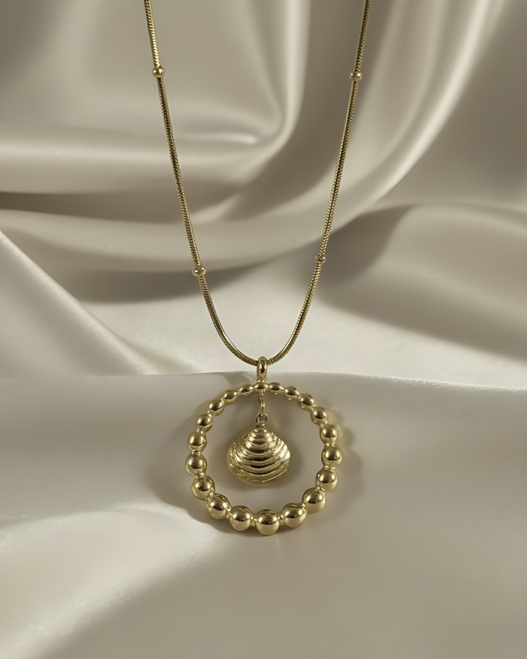 Gold Beaded Circle Pendant Necklace with Seashell Charm A gold necklace featuring a large beaded circle pendant with a smaller seashell charm suspended inside, hanging from a gold snake chain on white silk.