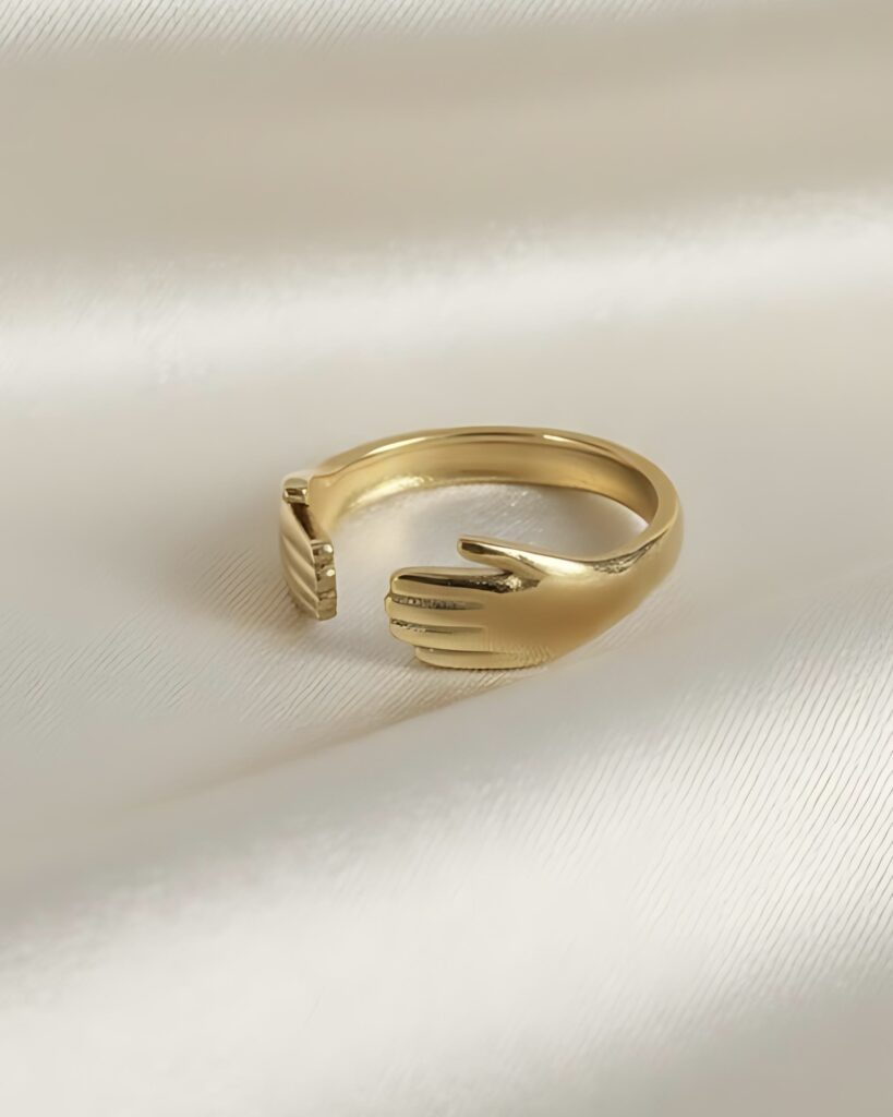 A gold open wrap ring with the ends shaped like two hands embracing the finger, creating a hug symbol, resting on white satin.