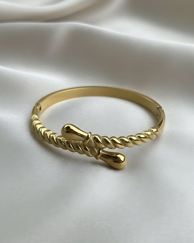 A wide gold hinged bangle bracelet featuring a central open area where the band is sculpted into a twisted rope design with teardrop ends, resting on white silk.