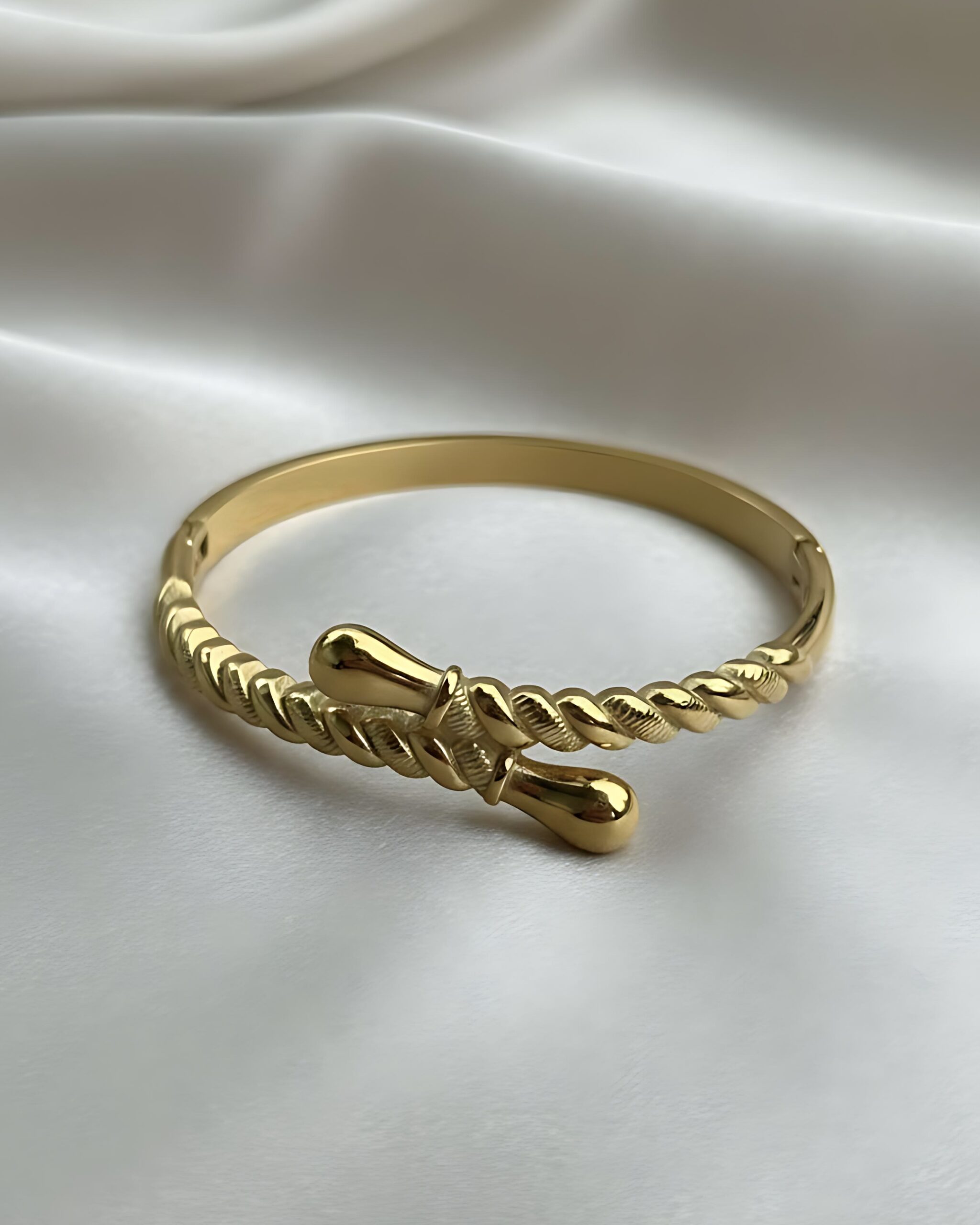 Gold Rope Twist Open Cuff Bangle with Hinged Clasp A wide gold hinged bangle bracelet featuring a central open area where the band is sculpted into a twisted rope design with teardrop ends, resting on white silk.