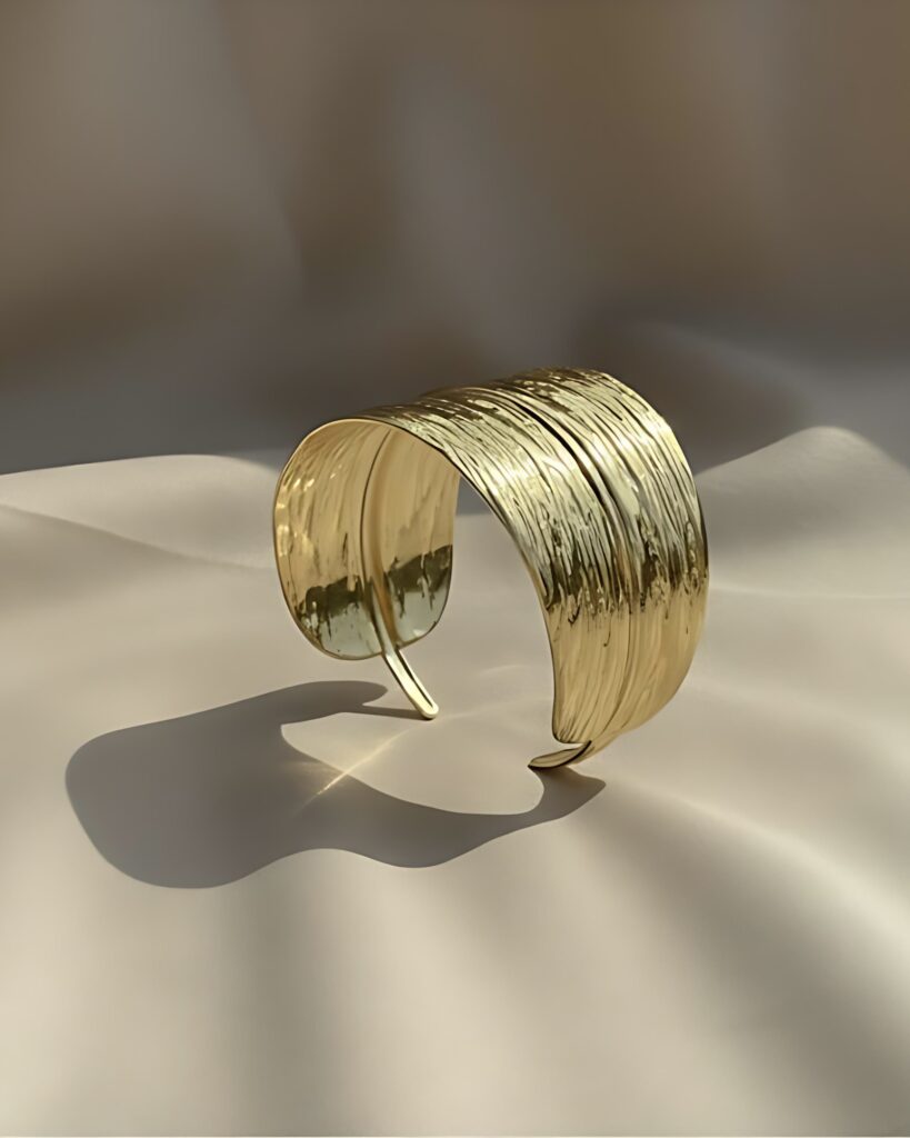 A wide gold open cuff bangle sculpted into a curved leaf or feather shape with a detailed, striated texture, resting on a beige satin background.