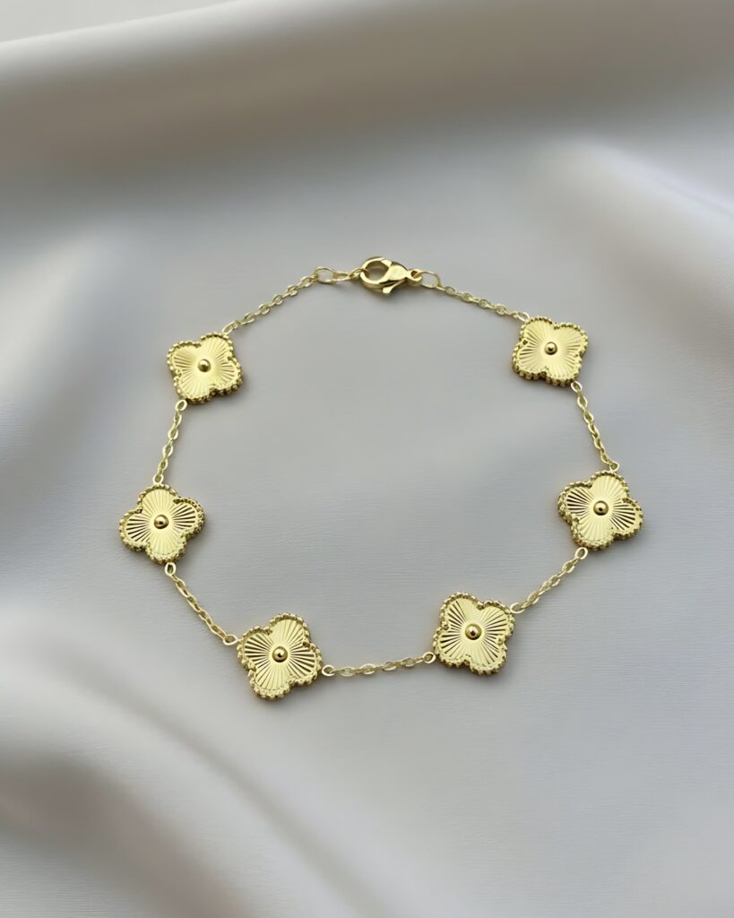 A gold link bracelet featuring five four-leaf clover charms with a subtle sunburst texture at the center of each, connected by a delicate chain, resting on white silk.