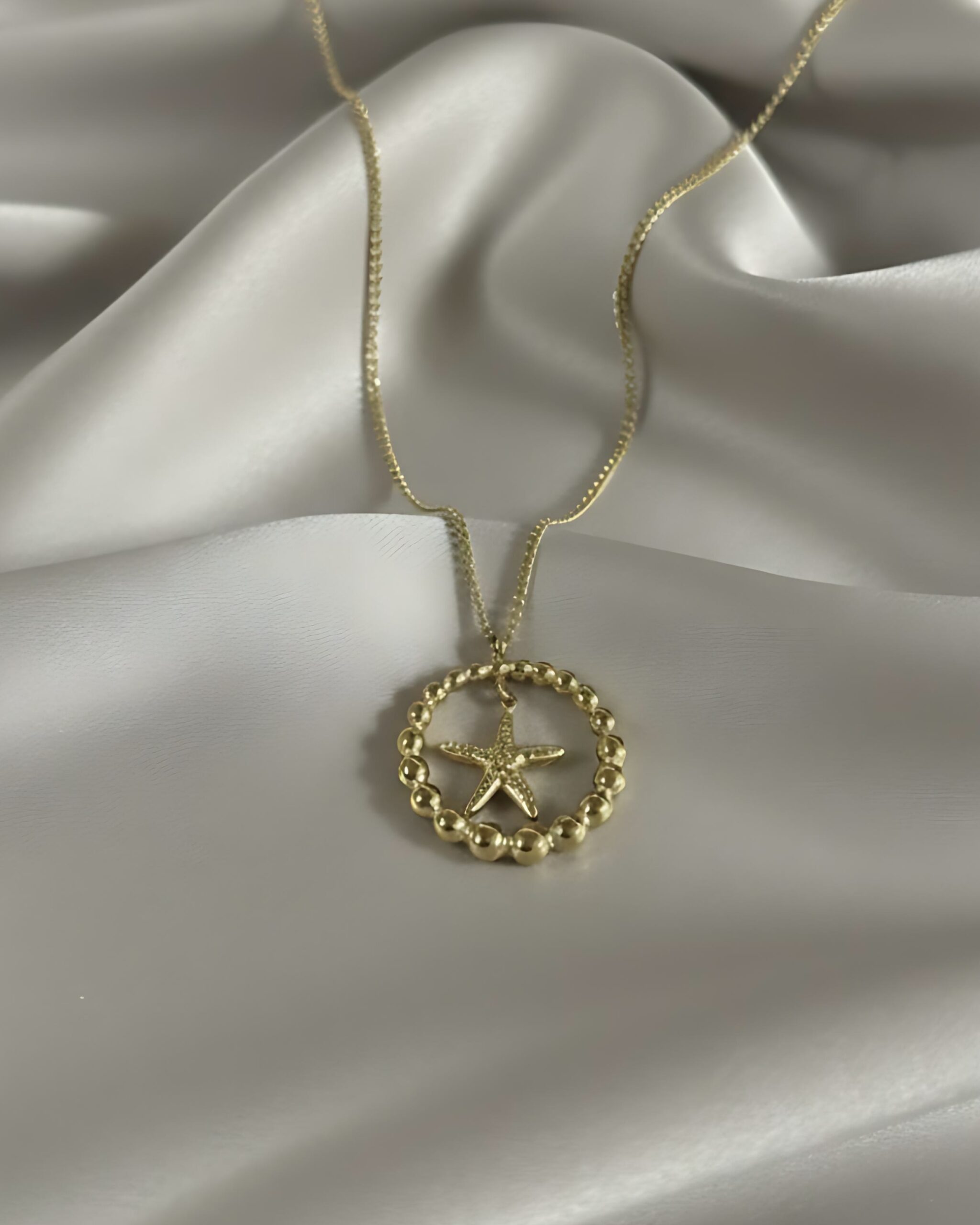 Gold Beaded Circle Pendant Necklace with Dainty Starfish Charm A gold necklace featuring a large beaded circle pendant with a smaller starfish charm suspended inside, hanging from a fine gold chain on white silk.