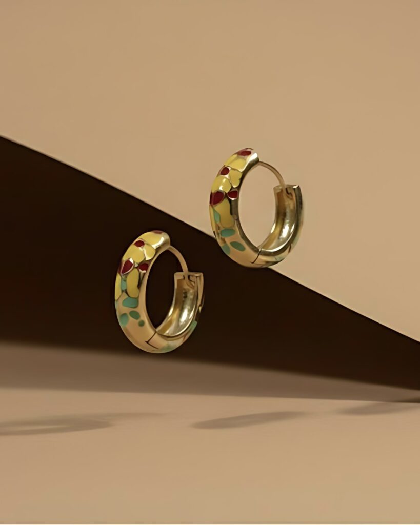 A pair of gold huggie hoop earrings featuring a continuous, hand-painted floral pattern of yellow sunflowers, red accents, and green enamel, set against a brown background.