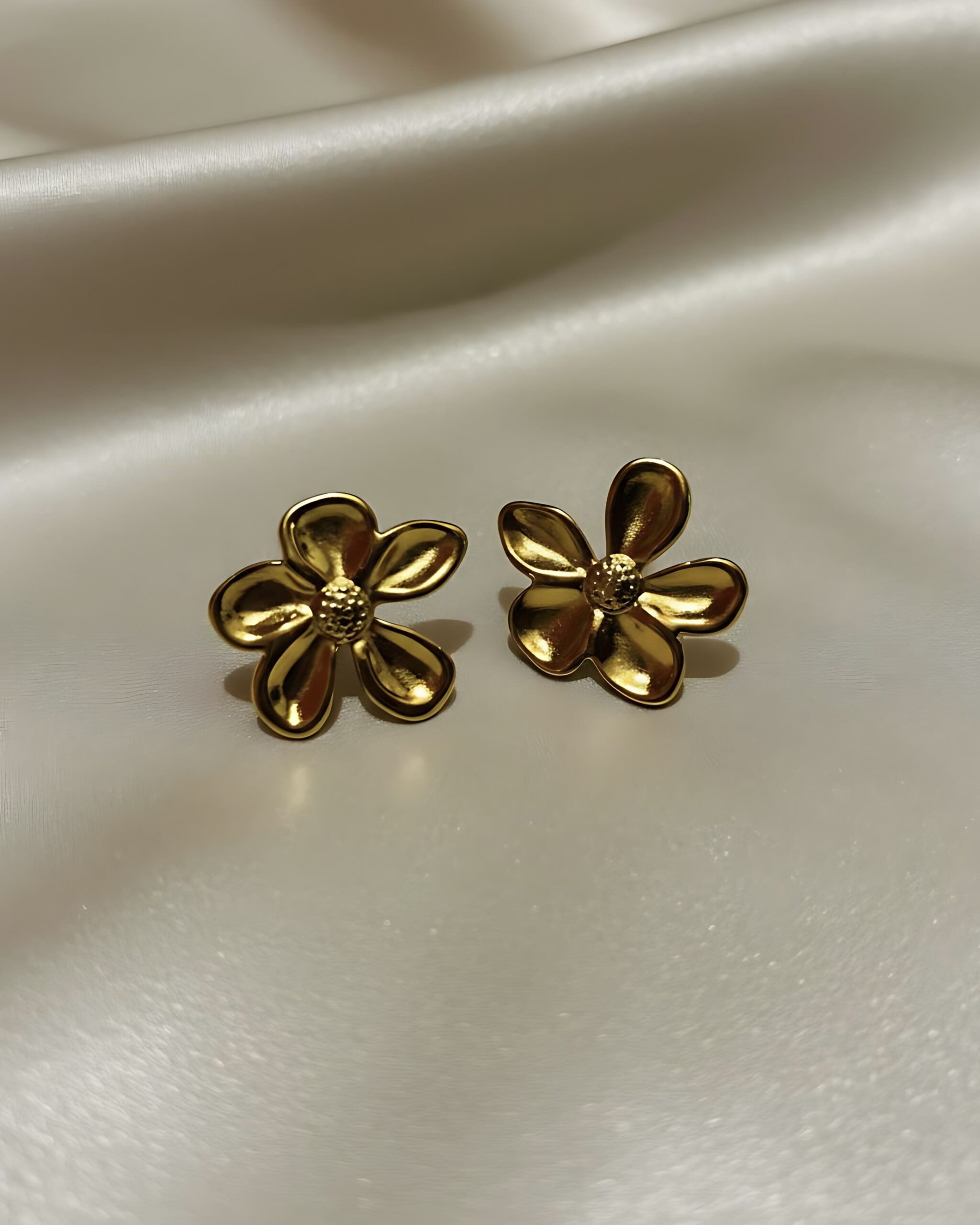 Minimalist Daisy Bloom Studs-Everyday Nature Inspired Jewellery A pair of small, gold-toned stud earrings sculpted into the shape of a five-petal flower with a textured center, resting on a white silk background.