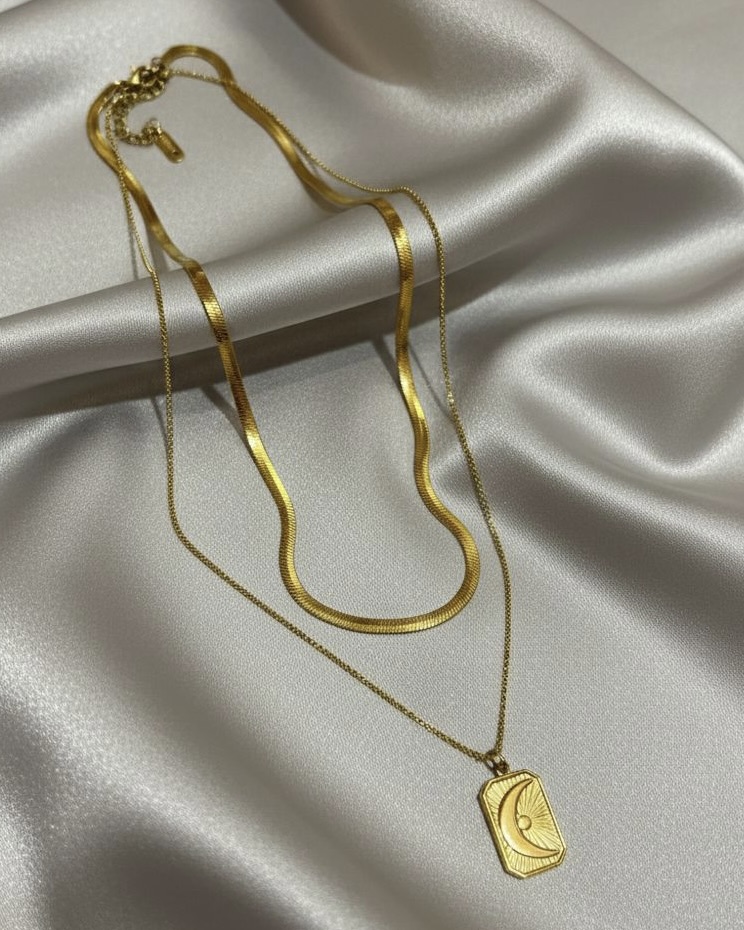 A gold pre-layered necklace set featuring a wide snake chain on top and a fine chain with a small rectangular moon pendant on the bottom, resting on white silk.