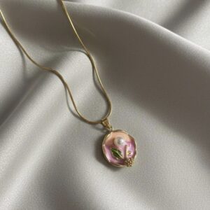 A gold snake chain necklace with a circular, scalloped pendant featuring a hand-painted pink and green enamel flower with a small pearl center, resting on white silk.