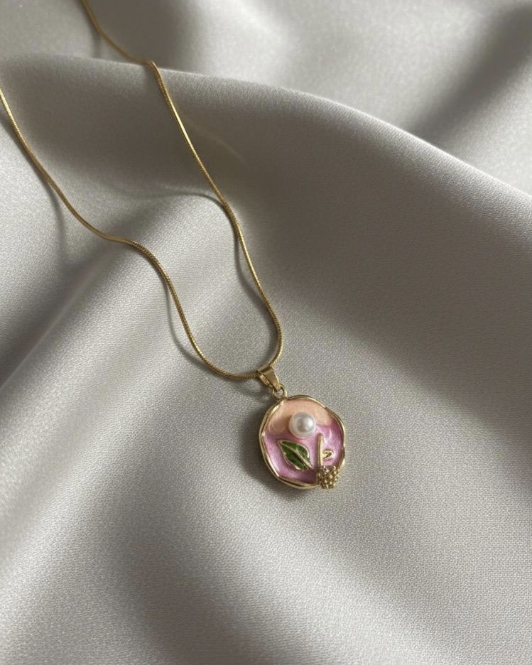 A gold snake chain necklace with a circular, scalloped pendant featuring a hand-painted pink and green enamel flower with a small pearl center, resting on white silk.