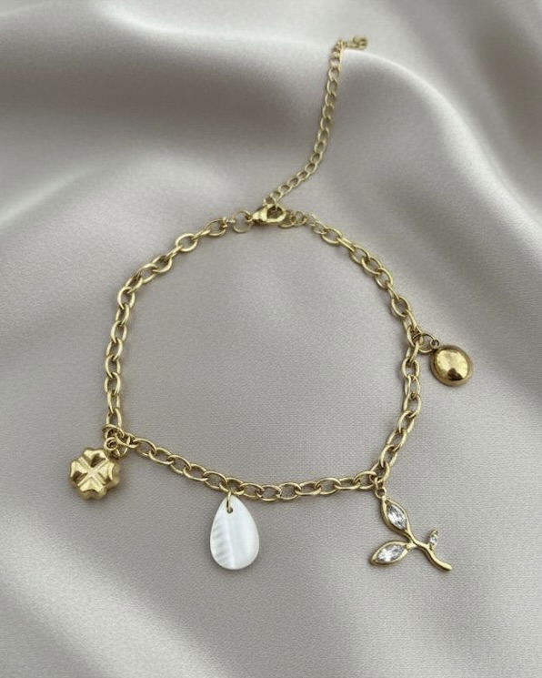 A gold link chain bracelet featuring multiple dangling charms, including a four-leaf clover, a mother-of-pearl teardrop, and a crystal-accented leaf, resting on white silk.