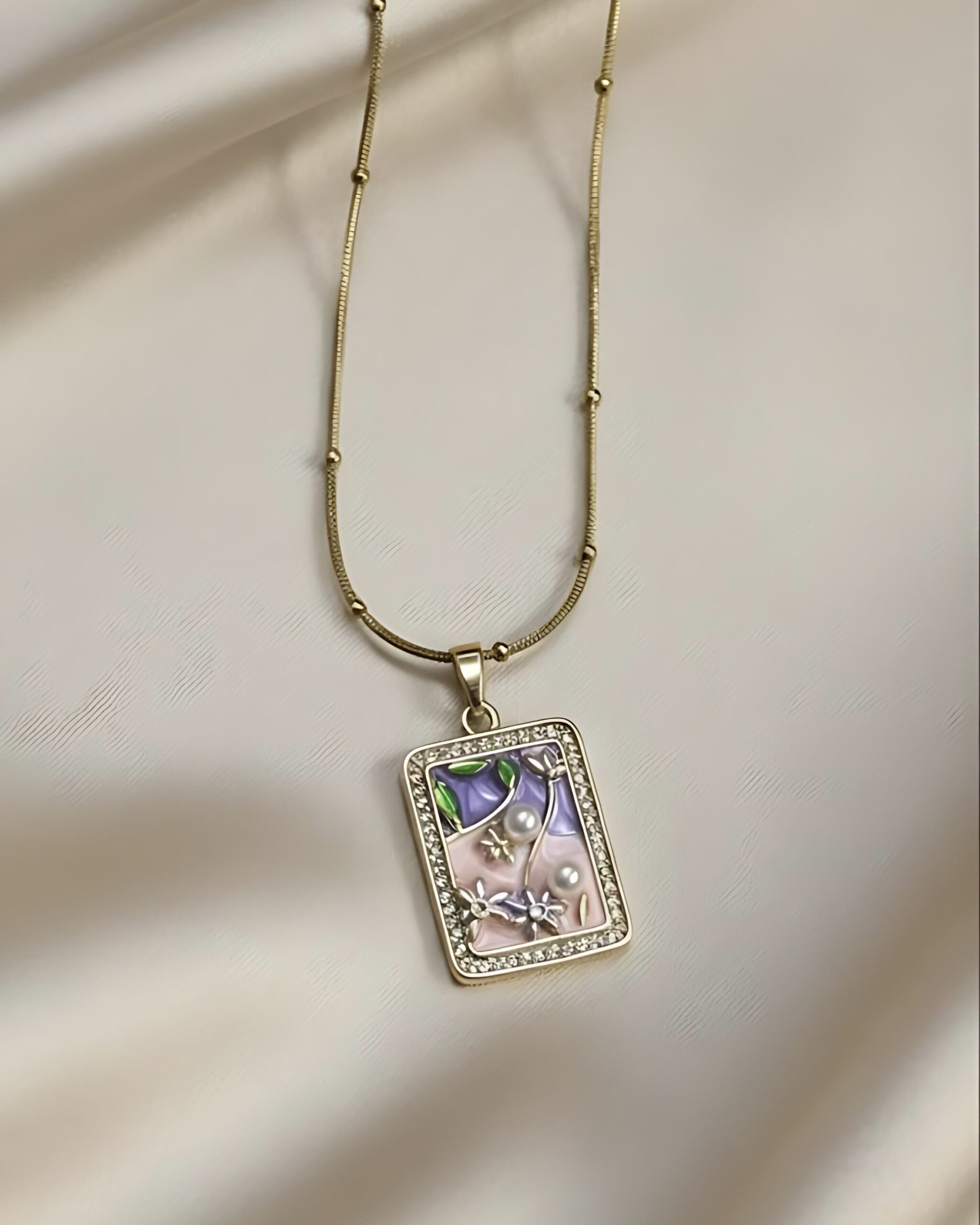 Gold Rectangle Pendant Necklace with Hand-Painted Enamel Flowers and Pave Border A gold satellite chain necklace with a small, rectangular pendant featuring a hand-painted floral enamel scene with tiny pearls and a sparkling micro pave crystal border, resting on white silk.