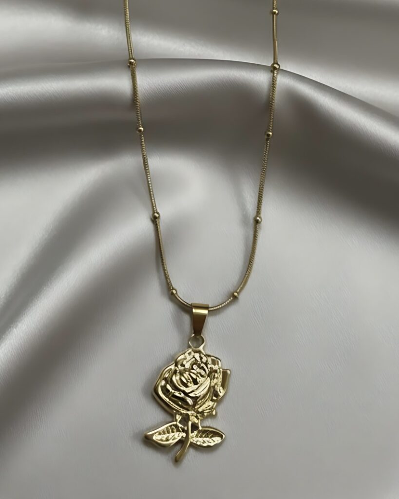 A gold necklace with a detailed, textured rose flower pendant suspended from a fine beaded satellite chain, resting on a white silk background.